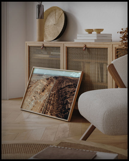 Ein gerahmtes Poster von einer Straße, die durch eine zerklüftete Landschaft in der Atacama-Wüste führt, an einer Holzkommode angelehnt und stehend auf dem Holzboden