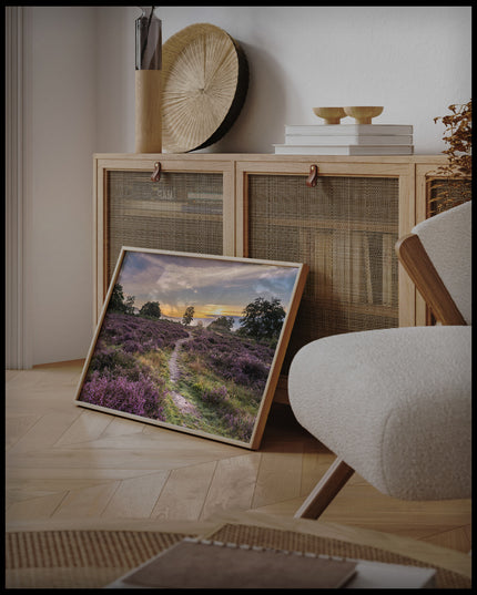 Ein gerahmtes Poster von einer Heidefläche mit lila Blumen im Sonnenuntergang, an einer Holzkommode angelehnt und stehend auf dem Holzboden