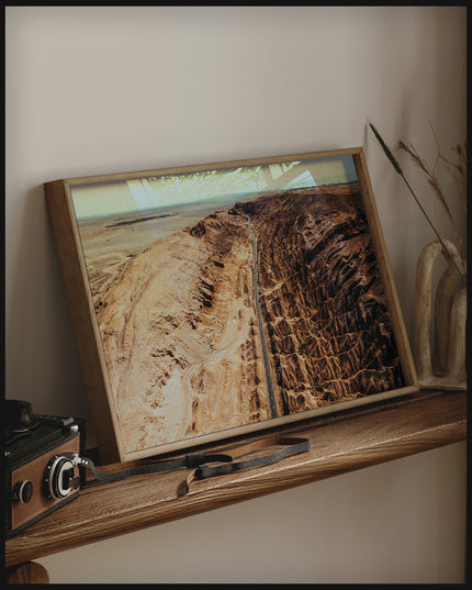 Ein gerahmtes Poster von einer Straße, die durch eine zerklüftete Landschaft in der Atacama-Wüste führt, auf einem Holzregal an die Wand angelehnt