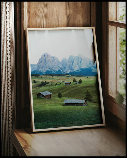 Ein gerahmtes Poster von einer grünen Alm mit kleinen Hütten und den Dolomiten im Hintergrund, vor einem Fenster an die Wand angelehnt