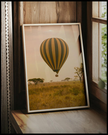 Ein gerahmtes Poster von einem Heißluftballon, der bei Sonnenuntergang über der afrikanischen Savanne schwebt, vor einem Fenster an die Wand angelehnt