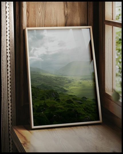 Ein gerahmtes Poster von einer grünen Landschaft in Irland mit Sonnenstrahlen, vor einem Fenster an die Wand angelehnt