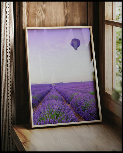 Ein gerahmtes Poster von einem Lavendelfeld und einem Heißluftballon mit lila-weißem Muster, der über dem Feld schwebt, vor einem Fenster an die Wand angelehnt