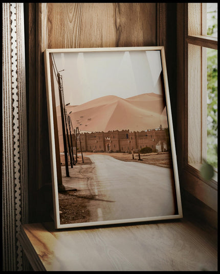 Ein gerahmtes Poster von einer Straße, die zu einer Siedlung am Rande einer weitläufigen Wüstenlandschaft mit hohen Sanddünen führt, vor einem Fenster an die Wand angelehnt