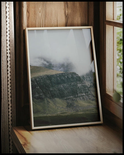 Ein gerahmtes Poster von einem steilen Abhang eines Berges, der von Gras bedeckt und von Nebel umschlungen ist, vor einem Fenster an die Wand angelehnt