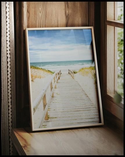 Ein gerahmtes Poster von einem Steg aus Holz der durch sandige Dünen zum Strand führt, vor einem Fenster an die Wand angelehnt