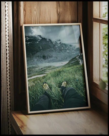 Ein gerahmtes Poster aus der Egoperspektive von einer Person die im Gras sitzt und auf eine Berglandschaft blickt, vor einem Fenster an die Wand angelehnt