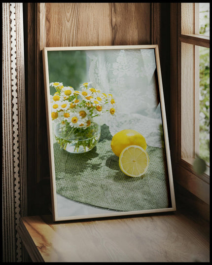 Ein gerahmtes Poster von zwei Zitronen und Blumen in einem Glas auf einer grünen Unterlage aus Stoff, vor einem Fenster an die Wand angelehnt