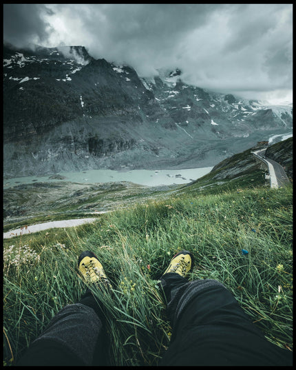 Ein Poster aus der Egoperspektive von einer Person die im Gras sitzt und auf eine Berglandschaft blickt.