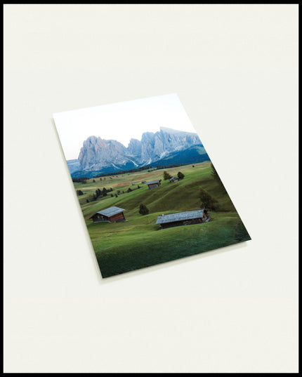 Eine Postkarte von einer grünen Alm mit kleinen Hütten und den Dolomiten im Hintergrund.